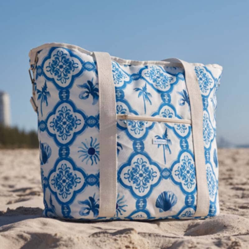 A blue and white patterned promotional products beach bag with cream-colored straps sits on sandy beach, with buildings, trees, and a blue sky in the background.