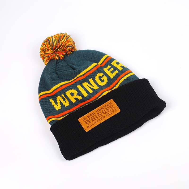 A knitted beanie hat with "WRINGER" in yellow letters, a multicolored pom-pom, and a black folded brim featuring a brown patch that reads "RED CENTER WRINGER BACKYARD ULTRA"—perfect custom merchandise for your promotional products lineup.