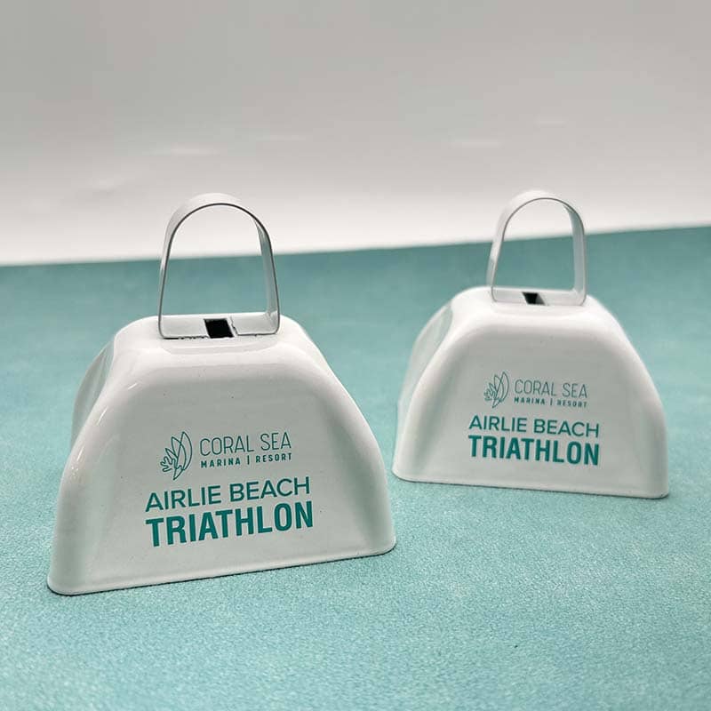 Two white cowbells with metal handles, featuring the "Coral Sea Marina Resort" logo and "Airlie Beach Triathlon" text, are displayed on a light turquoise surface—perfect custom merchandise for your next event or promotional products campaign.