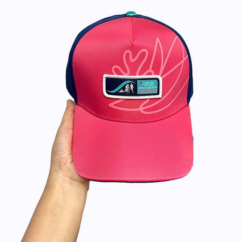 A hand holds a bright pink and navy blue baseball cap with abstract leaf designs and a rectangular patch featuring a wave, sun, and text—perfect as custom merchandise or eye-catching promotional products.