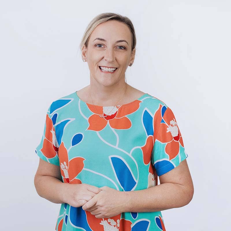 A woman with light hair pulled back, smiling, wearing a colorful floral top with red, orange, blue, and white patterns—perfect for custom merchandise—stands against a plain white background.