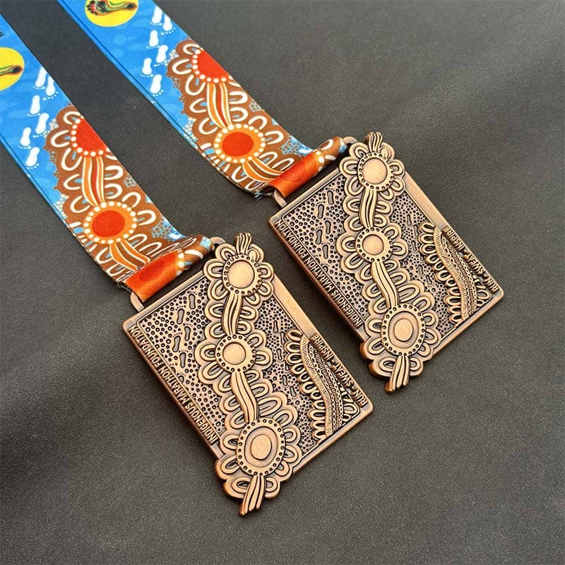 Two rectangular bronze medals with intricate, symmetrical patterns and floral designs—perfect as custom merchandise. The medals hang from blue ribbons decorated with orange suns and stylized white shapes against a plain dark background.