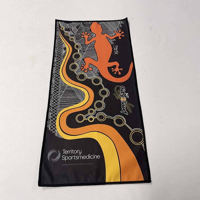 A rectangular towel features an orange and yellow abstract design with a gecko illustration, circular patterns, and the text "Territory Sportsmedicine" and "NT City2Surf"—perfect as custom merchandise or promotional products.