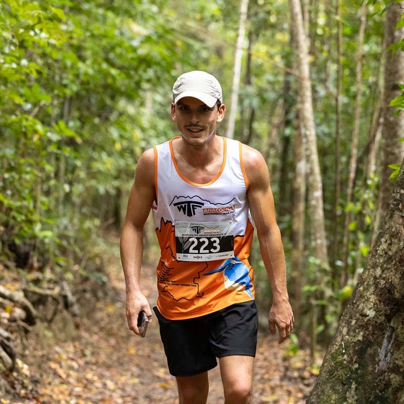 A man wearing a white cap, orange and white running tank top, and black shorts jogs on a forest trail surrounded by green trees. Sporting race bib 223, he carries custom merchandise in his right hand as part of the event's promotional products.