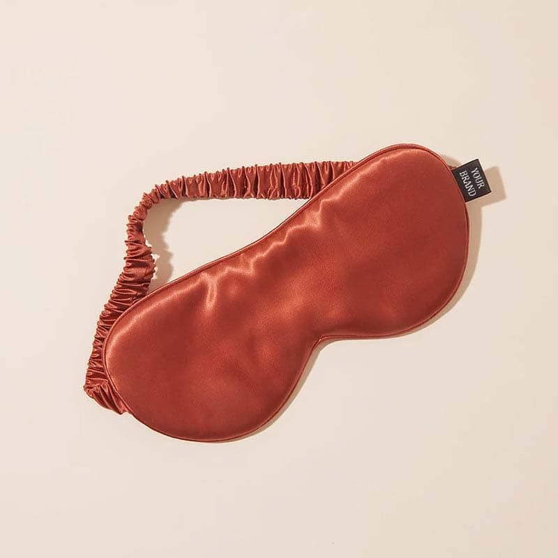 A shiny, rust-colored silk sleep mask with an elastic strap rests on a light beige surface. Perfect as custom merchandise or promotional products, it features a small black tag attached to one side of the mask.