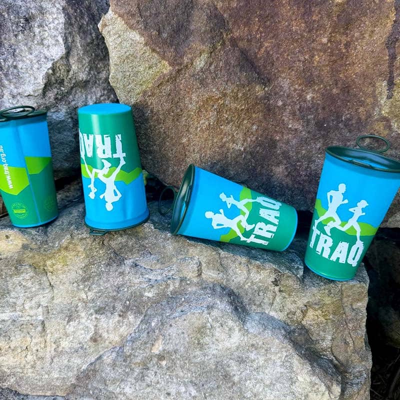 Three teal and green cans with silhouettes of people running and the word "TRAQ" are displayed on large, rough rocks; these eye-catching promotional products highlight unique custom merchandise for active lifestyles.