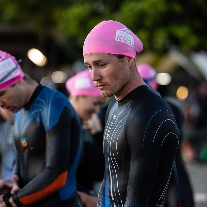 A triathlete in a black wetsuit and pink swim cap, featuring custom merchandise, stands among other competitors looking focused before the race. The outdoor background is slightly blurred, highlighting the anticipation at the start.