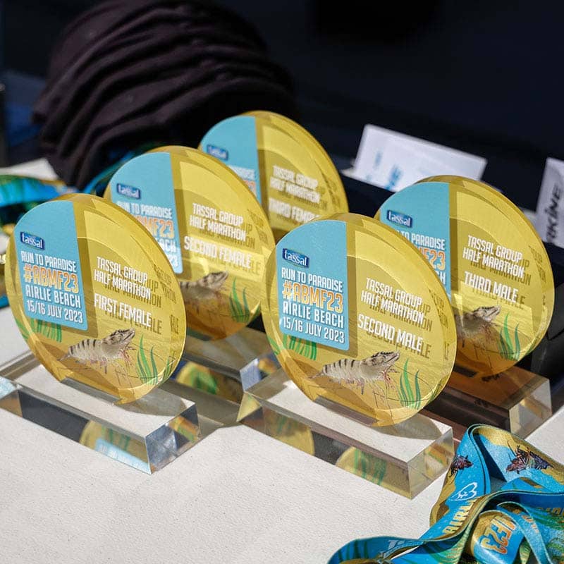 Five circular trophies with yellow and blue designs showcase awards for the "Run to Paradise" half marathon at Airlie Beach, 15–16 July 2023. Each trophy sits on a clear rectangular base—perfect custom merchandise for memorable events.