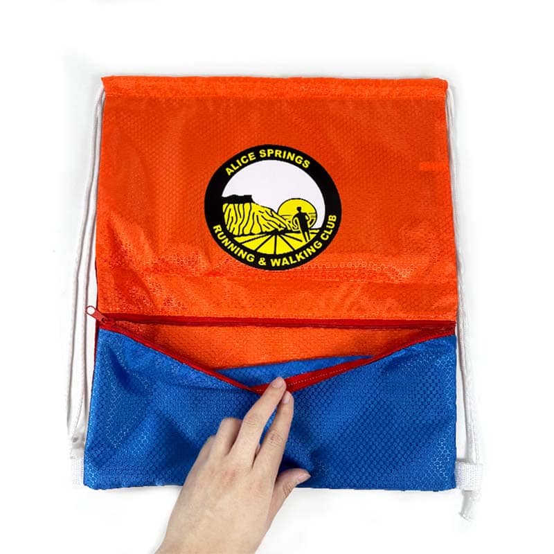 A hand opens the blue zippered pocket of a red and blue drawstring bag—custom merchandise for the "Alice Springs Running & Walking Club," featuring their zebra and sun logo. This eye-catching promotional product is set against a white background.