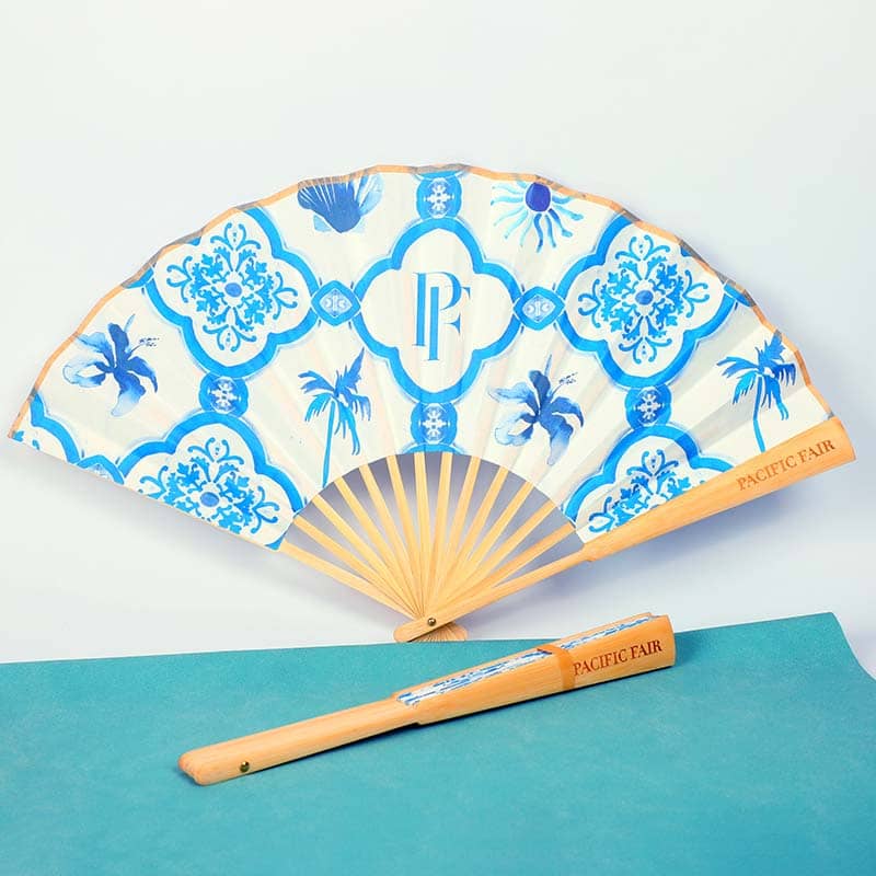 A decorative hand fan featuring blue floral and geometric patterns, wooden ribs, and "Pacific Fair" printed on the handle—an elegant piece of custom merchandise. The fan is displayed open, with its closed form placed below on a teal surface.