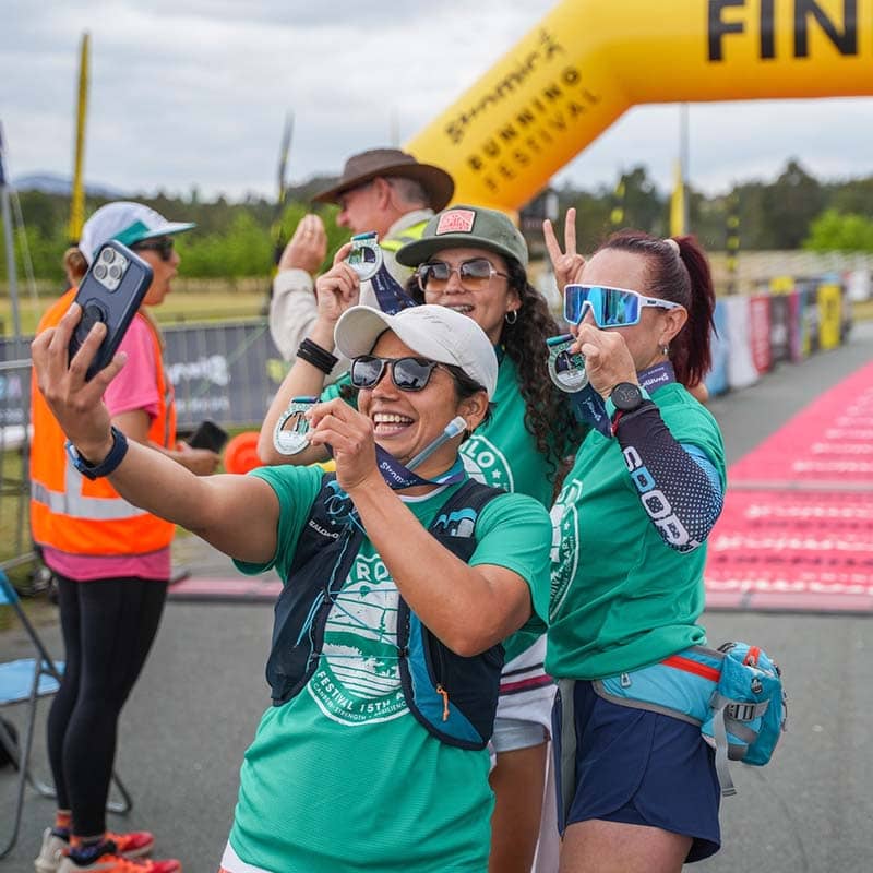 Three smiling women in green shirts pose for a selfie at a race finish line, showing off their medals and peace signs under a yellow “FINISH” arch—celebrating with custom merchandise and proud smiles.