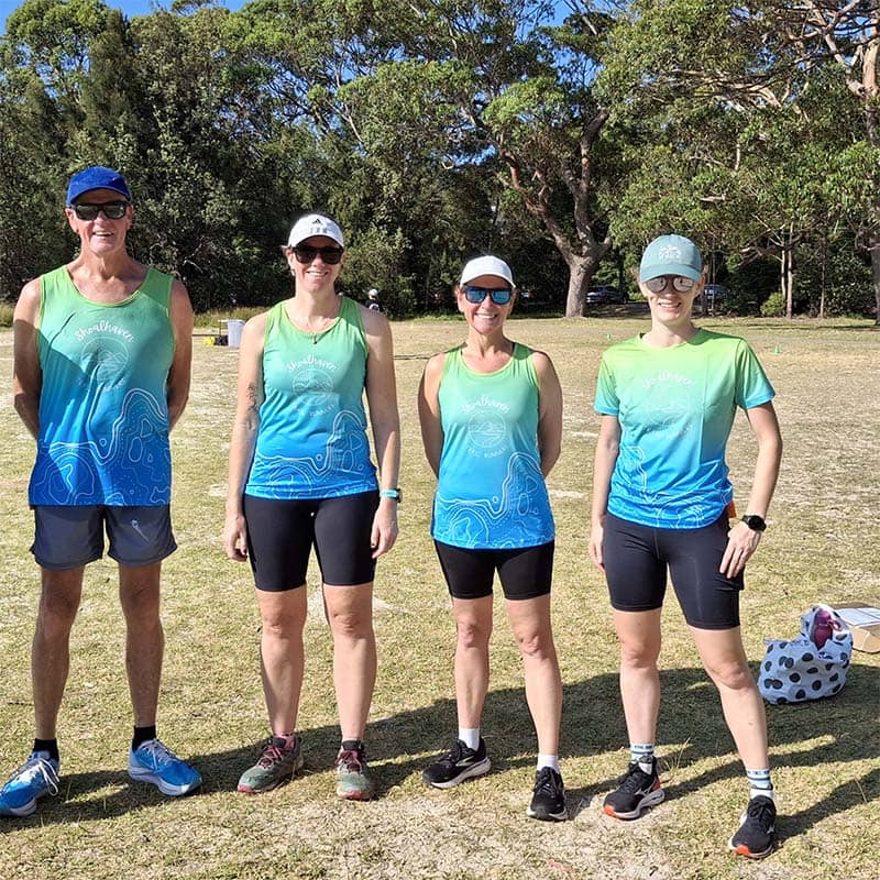 Four people stand outdoors on grass, all wearing matching blue-green athletic shirts and shorts, showcasing custom merchandise. Trees and sunlight are in the background as they appear ready for a sporting or running event.