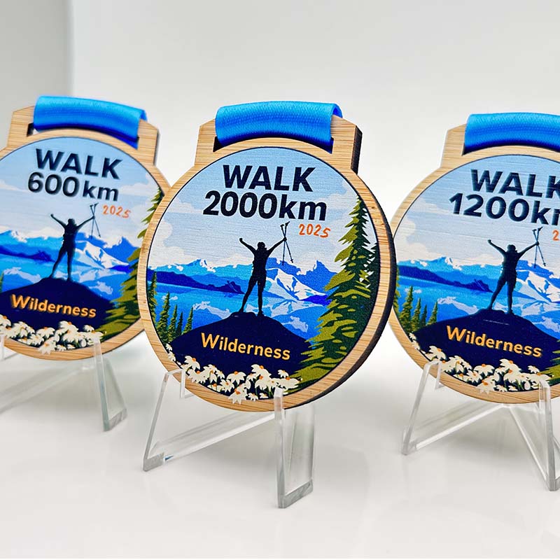 Three colorful medals on stands feature illustrations of a hiker with raised arms against a mountain backdrop, labeled "WALK 600km," "WALK 2000km," and "WALK 1200km." Perfect as custom merchandise or promotional products for Wilderness events.